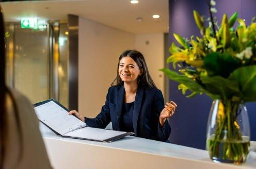 Smiling woman behind a reception desk, offering a sign in book. A vase of yellow flowers are on the desk in the foreground